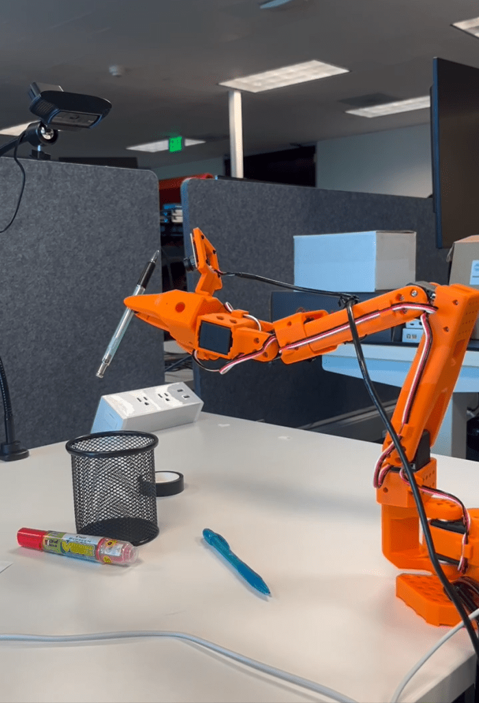 Orange robotic arm placing a pen into a black mesh holder on a desk.